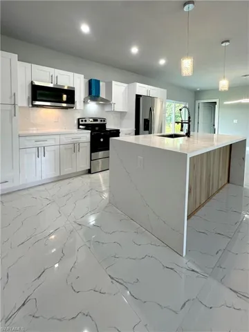 Kitchen with white cabinets, a center island with sink, decorative light fixtures, wall chimney range hood, and stainless steel appliances