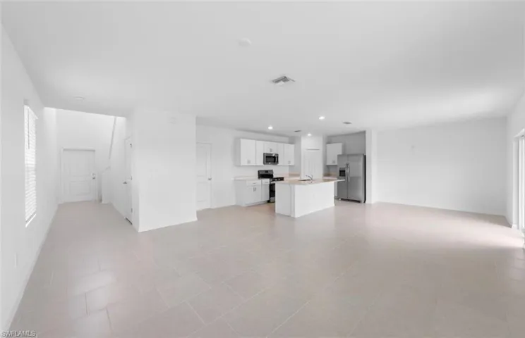 Expansive open concept living area with large format tile flooring throughout, flowing seamlessly into a well appointed kitchen featuring white shaker cabinets, a center island, stainless steel appliances and abundant natural light from multiple windows.