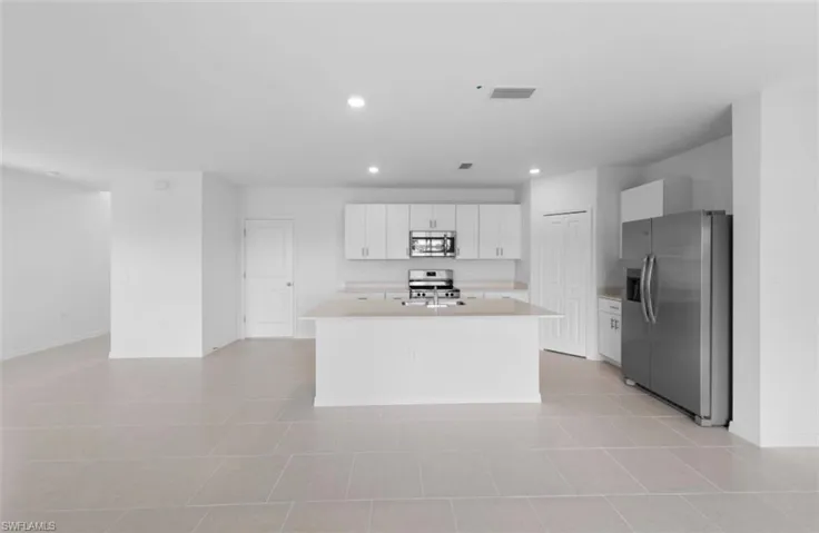 Open concept kitchen and living area showcasing a large center island with sink, white shaker cabinets, quartz countertops, stainless steel appliances and recessed lighting throughout — all set on large format tile flooring that flows seamlessly into the main living space.
