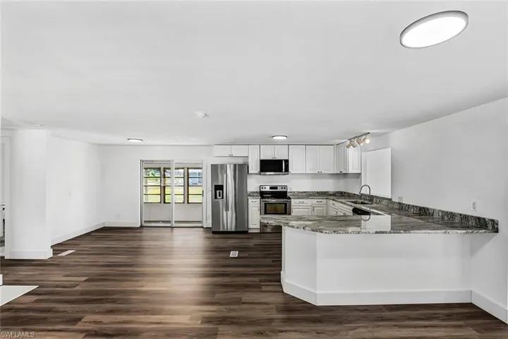 Expansive open-concept living area featuring wood-finish flooring, a contemporary kitchen with white cabinetry, stainless steel appliances, and a generously sized island with a stone countertop