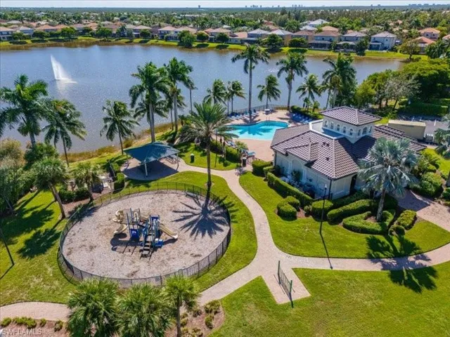 Community amenities featuring a lake with a fountain, a large swimming pool, a clubhouse with a tiled roof, a shaded playground, and paved walking paths surrounded by extensive landscaping and palm trees