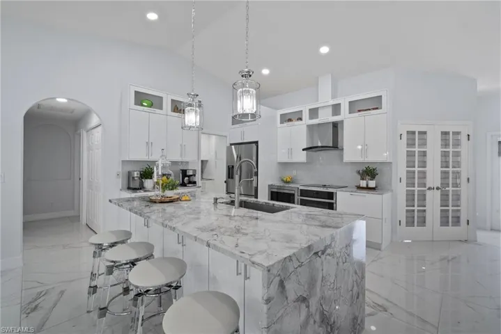 Kitchen featuring wall chimney range hood, light marble finish flooring, appliances with stainless steel finishes, lofted ceiling, and a kitchen bar