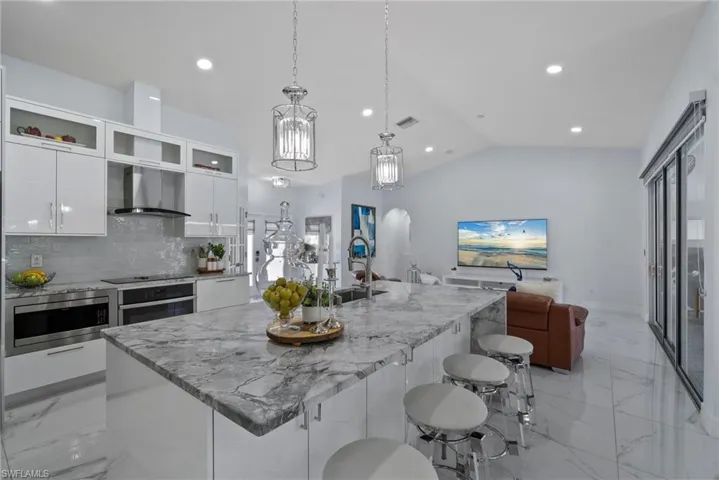 Kitchen featuring wall chimney exhaust hood, light marble finish flooring, vaulted ceiling, modern cabinets, and appliances with stainless steel finishes