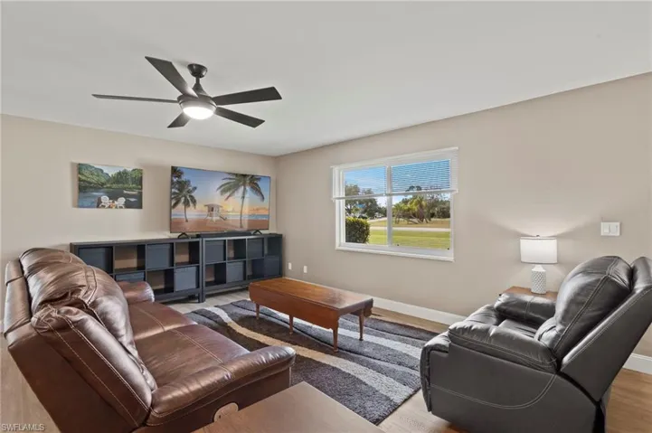 Natural light pours into the living room through the impact window with white blinds, illuminating the space and framing a serene view of greenery.
