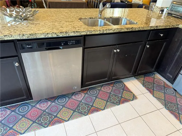 Kitchen featuring stainless steel dishwasher, dark cabinets, light tile patterned floors, and light stone countertops