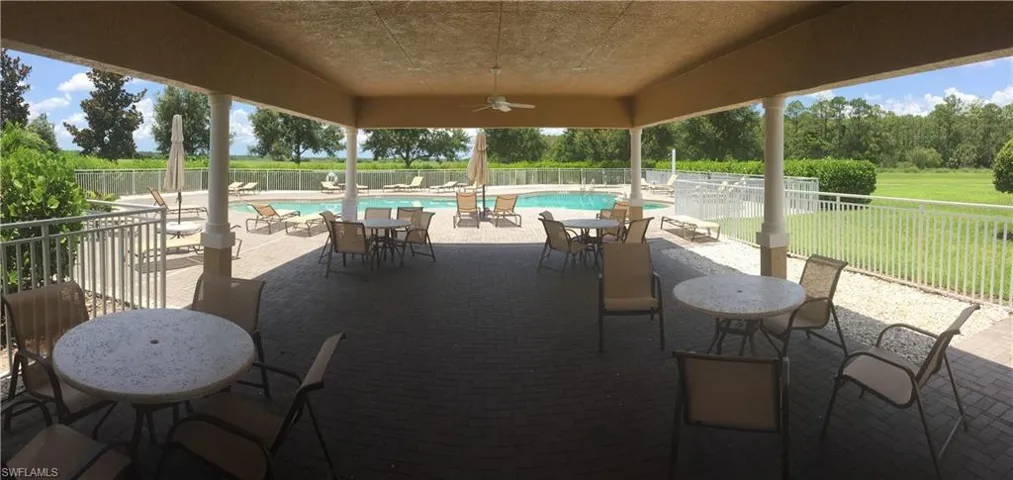Covered outdoor patio area featuring a ceiling fan, tables, and chairs, overlooking a swimming pool with lounge seating and surrounding green spaces