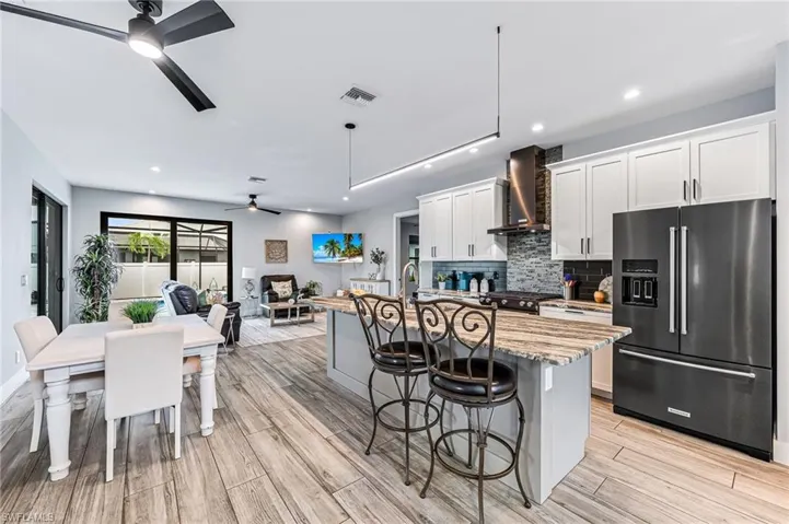 Kitchen with a breakfast bar area, a center island with sink, fridge with ice dispenser, ceiling fan, and wall chimney exhaust hood