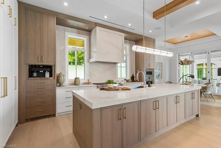 Kitchen with light wood-type flooring, a large island with sink, custom range hood, beamed ceiling, and light stone countertops