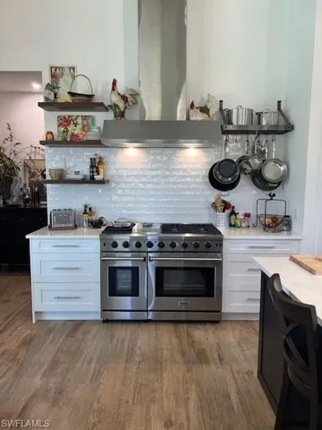 Kitchen featuring open shelves, white cabinets, range with two ovens, extractor fan, and light stone counters