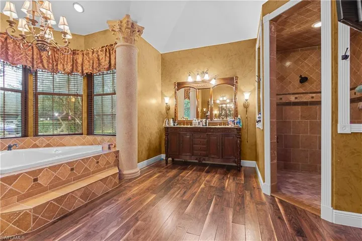 Spacious bathroom featuring a large soaking tub with tiled surround, an ornate column, a double vanity with framed mirrors, a walk-in shower with tiled walls and flooring, and wood-finish flooring