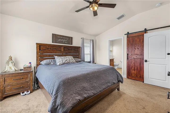 Bedroom with a barn door, ensuite bath, vaulted ceiling, light carpet, and ceiling fan
