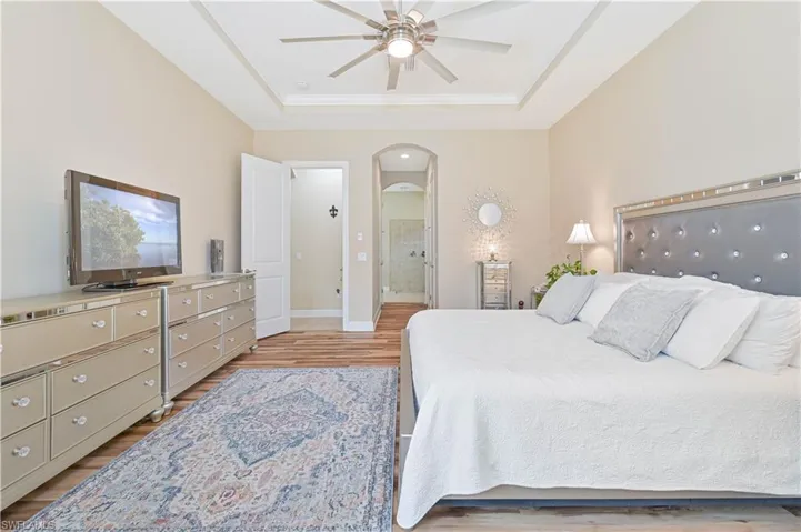 Bedroom featuring arched walkways, ensuite bath, light wood finished floors, a ceiling fan, and a tray ceiling