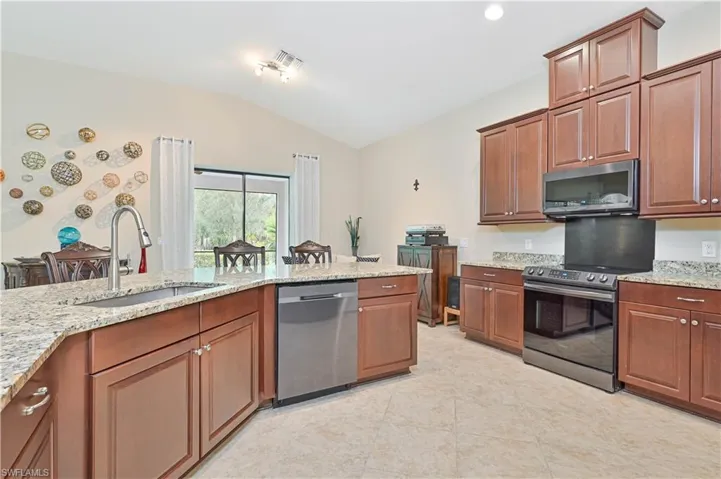 Kitchen featuring stainless steel appliances, brown cabinets, lofted ceiling, light stone countertops, and recessed lighting