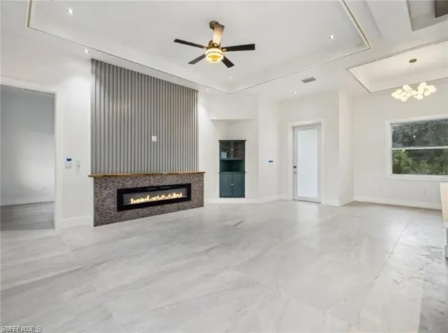 Unfurnished living room featuring baseboards, a glass covered fireplace, a tray ceiling, and ceiling fan with notable chandelier