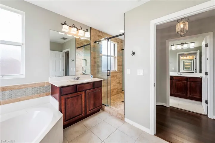 Bathroom featuring a corner soaking tub, a dark wood-finish vanity with a single sink and light-toned countertop, a glass-enclosed shower with natural stone-look tile, and ceramic tile flooring
