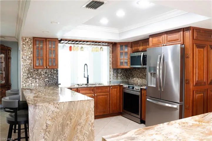 Kitchen featuring crown molding, brown cabinetry, glass insert cabinets, appliances with stainless steel finishes, and light stone counters