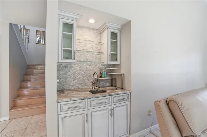 Bar with stairs, glass insert cabinets, backsplash, light stone countertops, and white cabinetry