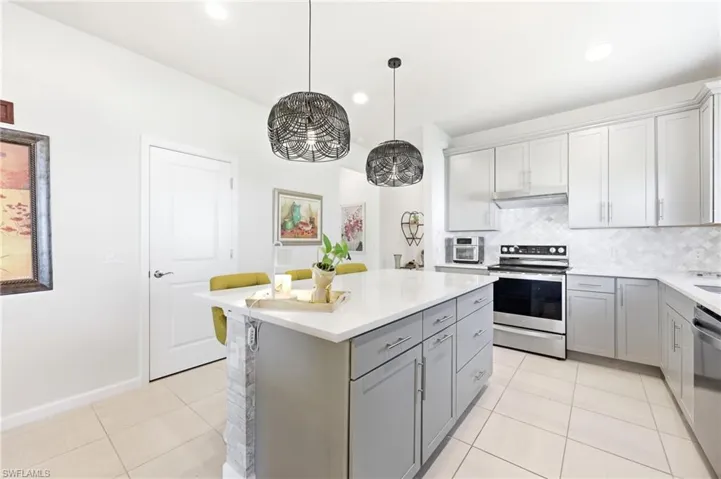 Modern kitchen featuring a central island with a light-toned countertop, light gray cabinetry, stainless steel appliances, a white tile backsplash, and ceramic tile flooring