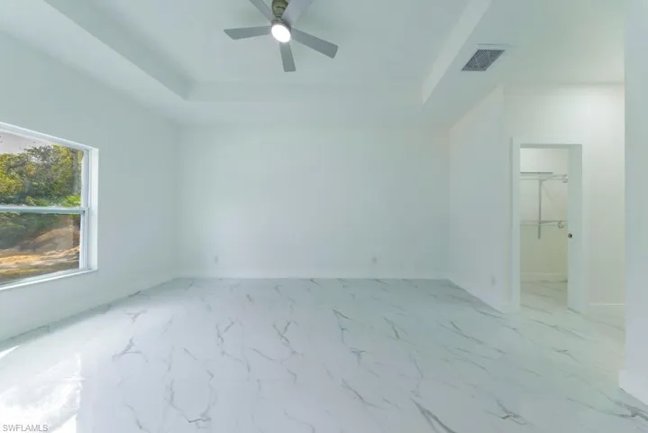 Unfurnished bedroom featuring a tray ceiling, light marble finish floors, a spacious closet, and ceiling fan