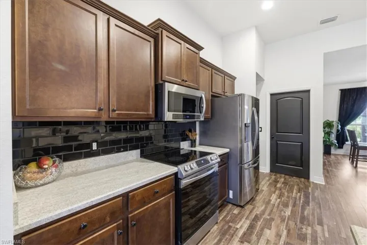 Kitchen with wood-finish flooring, dark wood cabinetry, light-toned countertops, a black subway tile backsplash, and stainless steel appliances