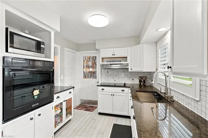 White cabinetry with black hardware, built-in oven and microwave, dark stone countertops, tile backsplash, and wood-finish flooring