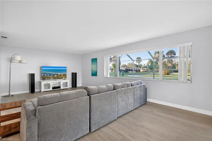 Bright interior space featuring light wood-finish flooring, a contemporary white media console, and a wide window offering views of mature landscaping