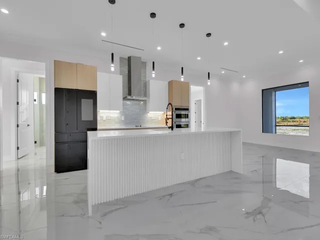 Kitchen featuring white custom wood cabinets, a center island with sink, wall chimney exhaust hood, and hanging light fixtures