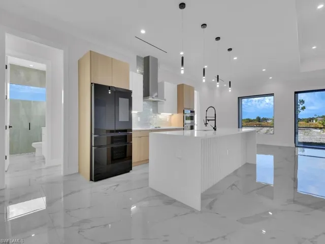 Kitchen featuring fridge, wall chimney exhaust hood, a kitchen island with sink, light brown and white custom wood cabinets, and hanging light fixtures