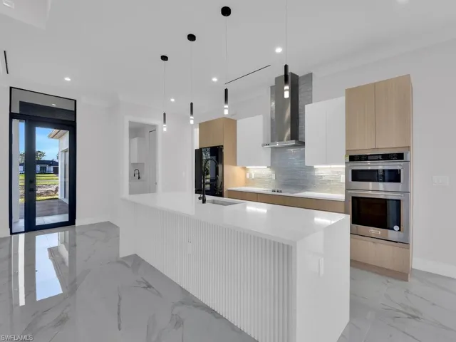 Kitchen with wall chimney exhaust hood, double wall oven, refrigerator,  a large island with sink, white custom wood cabinetry, and hanging light fixtures