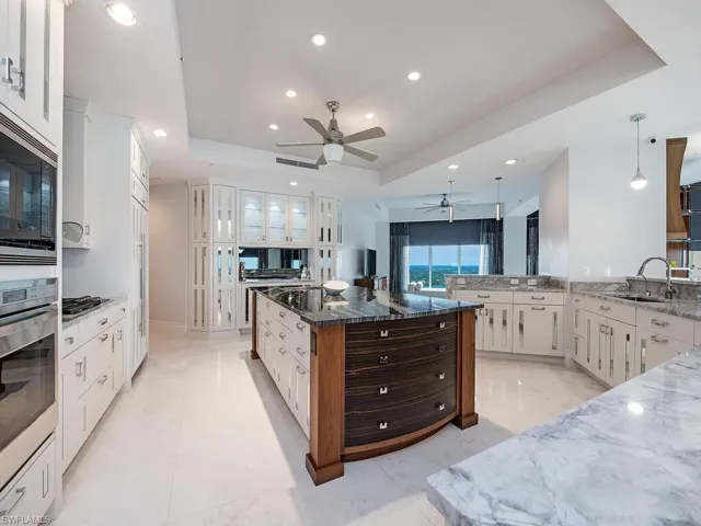 Kitchen with a raised ceiling, dark stone countertops, stainless steel appliances, a center island with sink, and white cabinets