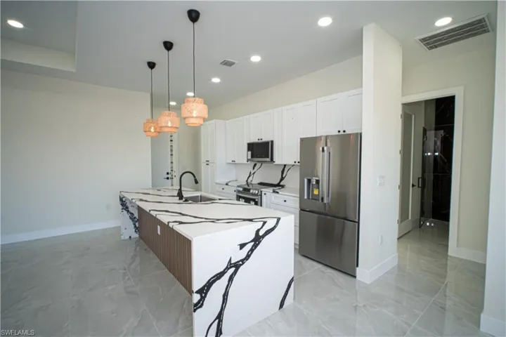 Kitchen with stainless steel appliances, an island with sink, white cabinetry, pendant lighting, and light marble finish floors