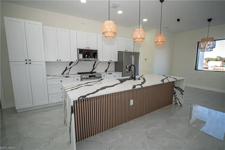 Kitchen with light stone counters, stainless steel appliances, a center island with sink, hanging light fixtures, and light marble finish floors