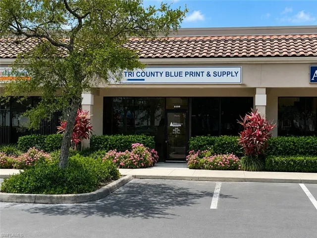 Commercial storefront featuring a prominent signage space, large front windows, a terracotta-style tile roof, stucco exterior, and landscaped beds with flowering plants and mature trees