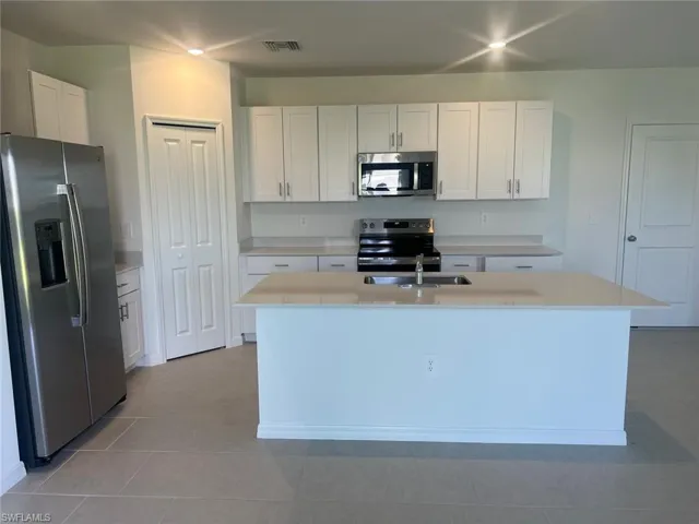 Kitchen featuring stainless steel appliances, white cabinets, an island with sink, light tile patterned floors, and light stone counters