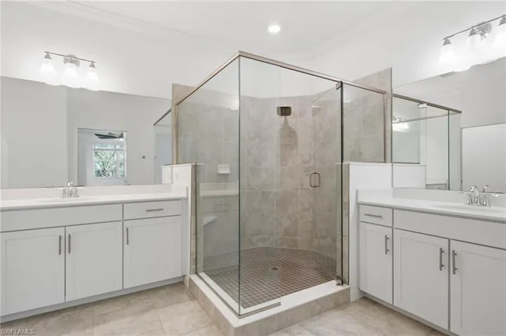 Dual white vanities with clean-lined cabinetry, glass-enclosed shower with tile surround, mosaic tile flooring, and integrated wall mirrors