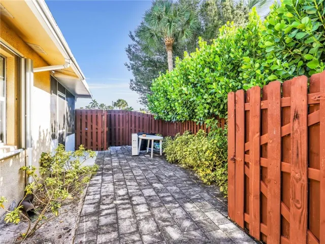 Paved outdoor area featuring a red-stained privacy fence, mature green foliage, and an adjacent building exterior with an awning