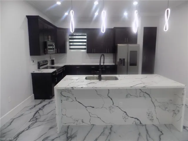 Kitchen featuring appliances with stainless steel finishes, ornamental molding, dark cabinetry, light stone counters, and a kitchen island with sink