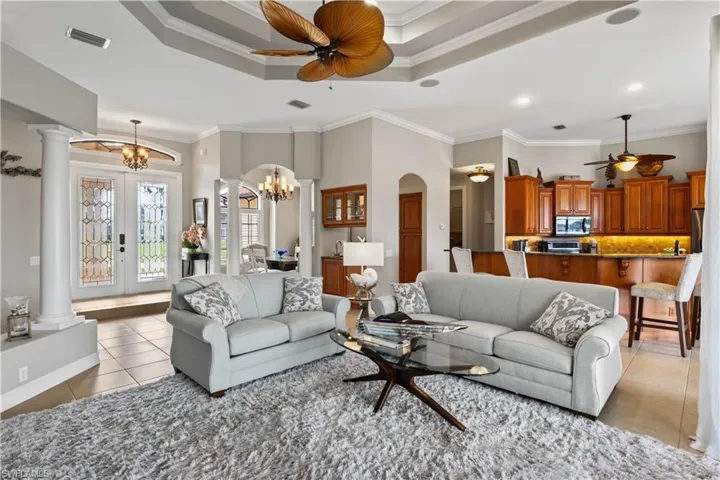 Tiled living room featuring a tray ceiling, crown molding, ceiling fan with notable chandelier, and decorative columns
