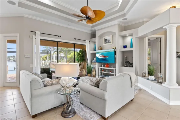 Tiled living room featuring ornamental molding, built in features, a raised ceiling, decorative columns, and ceiling fan