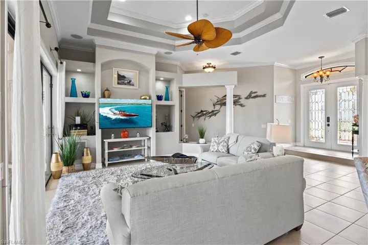 Tiled living room with ceiling fan with notable chandelier, built in features, crown molding, and a tray ceiling