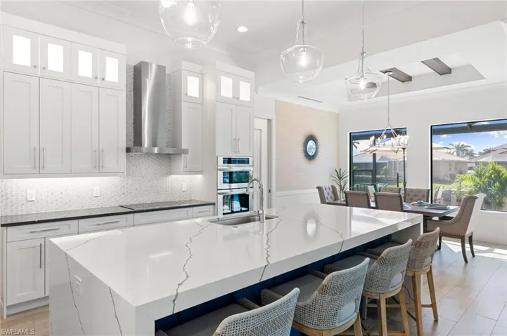 Kitchen featuring stainless steel double oven, white cabinetry, a sink, wall chimney range hood, and an island with sink