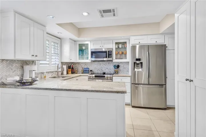 Kitchen with stainless steel appliances, a peninsula, light stone counters, glass fronted cabinets, and white cabinets