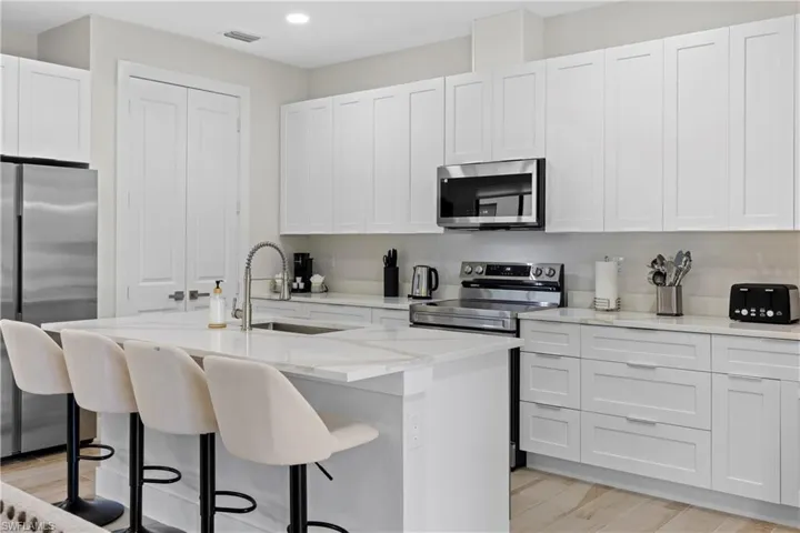 Contemporary kitchen featuring an island with integrated sink, stainless steel appliances, white shaker cabinetry, stone countertops, and wood-finish flooring