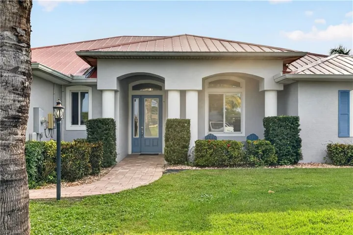 Stucco exterior with a metal roof, arched entryways, a brick paver pathway, and mature landscaping