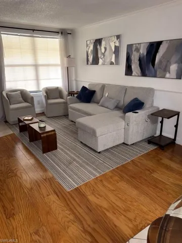 Living area featuring wood flooring, neutral wall tones, white crown molding, and a large window with blinds and sheer curtains