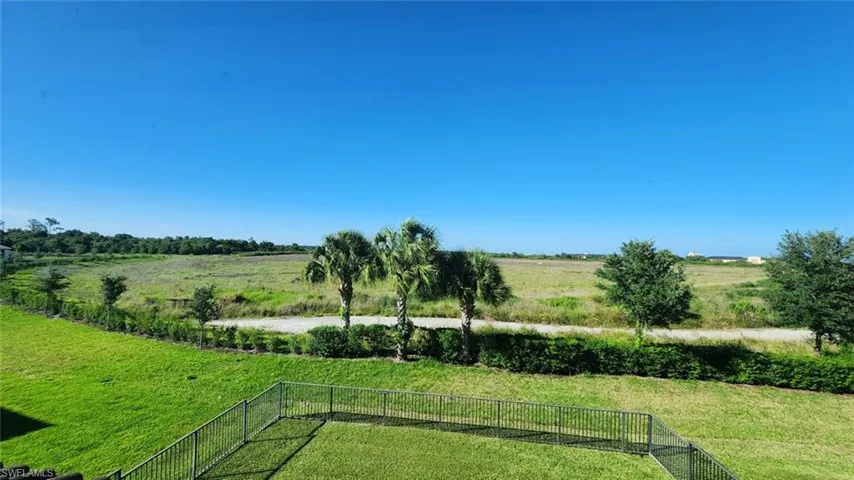 Expansive green lawn with a black metal fence, bordered by mature trees and a distant field under a clear blue sky