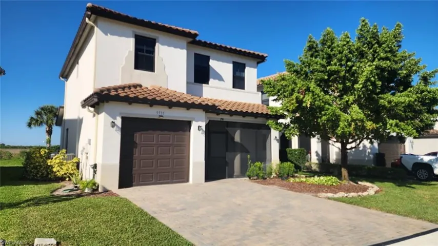 Stucco exterior with a tile roof, featuring a paver driveway, two-car garage, and landscaped front yard with mature trees