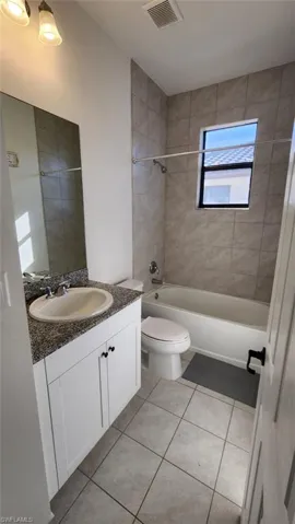 Bathroom featuring a vanity with a stone-finish countertop, a single basin sink, white cabinetry with black hardware, a bathtub with a tiled surround, and tiled flooring