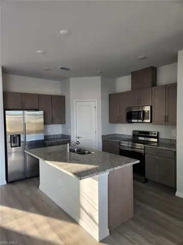 Kitchen featuring stainless steel appliances, dark wood-type flooring, dark stone counters, and a kitchen island with sink