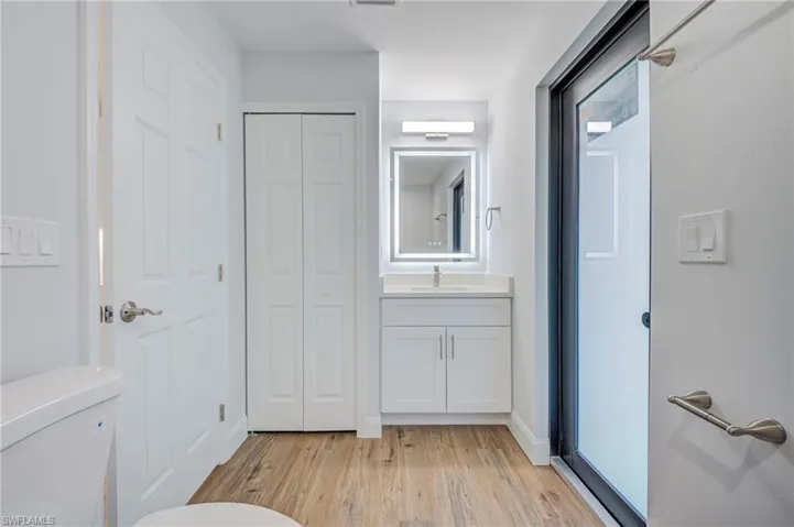Bathroom featuring a white vanity cabinet with a solid-surface countertop, an integrated sink, a rectangular mirror with integrated LED perimeter lighting, wood-finish flooring, and a floor-to-ceiling bi-fold closet